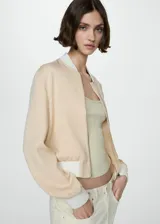 Chaqueta tipo bomber color beige, con cierre frontal de cremallera metálica, cuello, puños y ruedo en tejido de canalé blanco. Cuenta con dos bolsillos laterales de ribete.