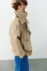 Trench corto oversize color beige con cuello subido de efecto doble, cierre cruzado con botones oscuros y bolsillos de vivo delanteros.