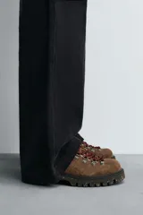 Zapato de cordones estilo trekking para hombre, confeccionado en piel con acabado de serraje color marrón. Presenta enganches metálicos para los cordones, cuello acolchado en color negro y una suela de goma con volumen y efecto moteado.