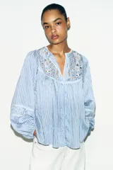 Blusa de rayas verticales blancas y celestes, con cuello redondo y escote en pico. Presenta detalles de bordados perforados y puntillas en el pecho y las mangas. Tiene mangas largas abullonadas con puños elásticos y cierre frontal con botones forrados.