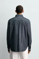 Camisa de corte relajado confeccionada en tejido de algodón denim, con cuello solapa, manga larga con puños abotonados y bolsillo plastrón en el pecho. Presenta cierre frontal de botones y bajo con aberturas laterales.