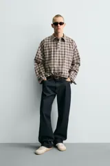 Camisa polo de corte boxy fit confeccionada en tejido de algodón con elasticidad, presenta un estampado de cuadros en tonos marrones, azules y blancos. Cuenta con cuello solapa, cierre frontal de botones, manga larga con puños abotonados y bajo con aberturas laterales.