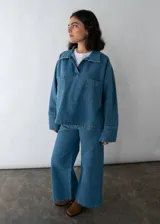 Camisa de denim azul con cuello polo, cierre frontal parcial y dos bolsillos cuadrados en el frente. Presenta un corte holgado y mangas largas.