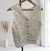 Chaleco de tejido de punto color crudo, con cuello en V y cierre frontal de botones marrones. Presenta un patrón calado con flores bordadas con lentejuelas plateadas.