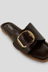 Sandalias Daniel Cassin de símil cuero color marrón oscuro, con una tira ancha cruzada en el empeine y una hebilla metálica rectangular dorada. Presentan puntera cuadrada y suela plana.