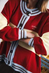 Cardigan oversized de punto grueso, color azul marino y blanco con franjas rojas y rosadas en el cuello, puños y solapa. Tiene botones grandes marrones y el logo de Anthea bordado en el pecho.
