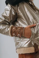 Campera puffer metalizada en tono dorado, con interior y detalles en corderito beige. Presenta capucha negra de corderito, cierre frontal y puños ajustables con tiras de cuero y tachas metálicas.