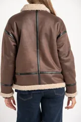 Campera tipo gamulán de corte oversize, color marrón con interior y cuello de corderito sintético en tono crema. Presenta detalles de ribetes en cuero sintético negro a lo largo de la prenda, cierre frontal metálico y puños tejidos.