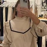 Blusa de lino color beige con cuello alto y volados en el frente y mangas. Presenta detalles de ribetes en terciopelo negro que contrastan con la tela principal. Posee mangas largas con puños abotonados y un diseño romántico y femenino.