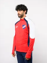 Buzo de entrenamiento Umbro del Club Nacional de Football, con cierre frontal parcial y cuello alto. Presenta un diseño con bloques de color azul, blanco y rojo, con panel frontal acolchado y mangas largas con aberturas para pulgares.