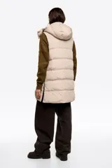 Chaleco acolchado de largo medio y corte en línea A, confeccionado en tejido técnico color beige con detalles en negro. Presenta cuello subido, capucha desmontable, cierre frontal de cremallera, bolsillos laterales con cremallera, logo bordado en el pecho y aberturas laterales con cremallera.