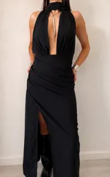 Vestido largo negro con escote halter profundo y detalle de lazo al cuello. Presenta un diseño drapeado en la cintura y una abertura lateral pronunciada en la falda.