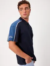 Remera polo gris melange de ajuste holgado, con cuello y cierre frontal de botones. Presenta detalles en azul marino y blanco en los hombros y mangas, y el logo de la marca bordado en el pecho.