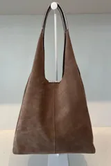 Bolso tipo tote de gamuza en color verde oliva, con diseño minimalista y asa integrada. Presenta una placa metálica con el logo de la marca en la parte inferior central y detalles de textura en el asa.