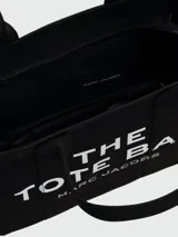 Bolso tipo tote de Marc Jacobs, confeccionado en lona resistente de color negro. Presenta el nombre del modelo estampado en blanco en la parte frontal y el logo de la marca debajo. Cuenta con doble asa superior para llevar al hombro o en la mano.