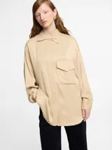 Camisa color beige de tejido satinado, corte oversize, manga larga, tapeta con botones invisibles y logo bordado en la espalda.