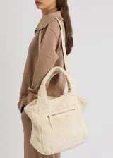 Bolso tote de sherpa color crema, con asas de mano y correa bandolera ajustable. Cuenta con cierre de cremallera y bolsillos internos.
