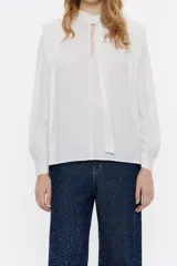 Blusa blanca de corte recto con cuello con abertura y lazo anudado. Presenta mangas largas con volumen y puños abotonados, y detalles de pliegues en los hombros.