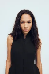 Chaleco entallado de color negro con cuello alto y cierre frontal con cremallera oculta. Presenta un diseño sin mangas con costuras verticales que definen la silueta y un bajo con ligero volumen acampanado.