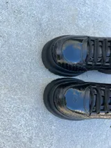 Zapato mocasín negro de cuero charol con diseño tipo reptil, cordones y suela track.