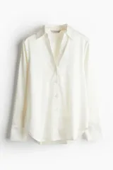 Blusa color blanco hueso de tela con brillo, cuello clásico, escote en V con botones, mangas largas con puños sencillos y bajo redondeado.