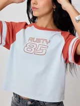 Remera corta de mangas ranglán color ladrillo con dos rayas celestes en los puños. Cuerpo celeste con estampado frontal "Rusty 85" en color ladrillo.