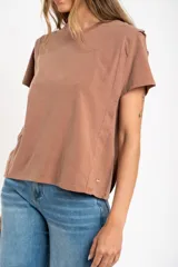 Remera de algodón color mostaza/naranja quemado, con cuello redondo y mangas cortas. Presenta un diseño con costuras externas visibles en los hombros y laterales, dándole un aspecto desestructurado.