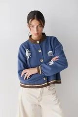 Cardigan de punto color crema con cuello redondo y cierre frontal de botones. Presenta detalles bordados de motivos celestiales y marinos, con terminaciones en tejido acanalado a rayas azules en cuello, puños y ruedo.