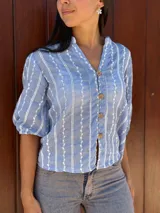 Camisa de corte recto y calce holgado, confeccionada en tela liviana color celeste con estampado de rayas verticales blancas con detalles de hojas. Presenta cuello en V, cierre frontal con botones de madera y mangas tres cuartos con terminación elástica.
