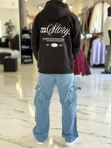 Canguro oversize negro con capucha y bolsillo tipo canguro. Presenta un gran estampado en la espalda con la inscripción "Own Your Story", texto descriptivo y un gráfico de globo terráqueo junto al año "2021".