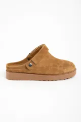 Zueco tipo mule de gamuza color camel, con forro interior de corderito visible en el borde. Presenta una tira ajustable con broche metálico en el lateral y suela de goma gruesa y dentada.