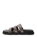 Sandalias planas Miss Carol modelo Nevada, color marrón, con tres tiras en el empeine decoradas con tachas metálicas plateadas. Una de las tiras cuenta con una hebilla de ajuste lateral. Poseen suela y plataforma baja de color negro.