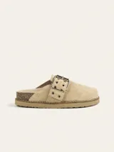 Zueco de gamuza color beige, con diseño de punta cerrada y talón descubierto. Presenta una tira ajustable sobre el empeine con hebilla metálica y ojales decorativos. Cuenta con una suela de corcho y base de goma antideslizante.