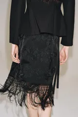 Falda corta de tiro medio, color negro, con diseño cruzado estilo pareo ajustable con lazada lateral. Cuenta con bordados tonales de motivos florales y bajo acabado con flecos largos.