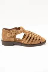 Sandalias cangrejeras de gamuza color visón, con diseño de tiras entrelazadas, tira ajustable con hebilla metálica en el tobillo y suela plana de goma marrón.