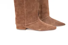 Bota de caña alta confeccionada en cuero gamuza color marrón, con diseño de solapa superior y detalle de hebilla metálica lateral. Presenta punta fina y suela plana.