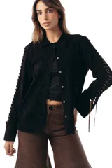 Camisa manga larga de gamuza ovina negra, con cuello clásico y cierre frontal de broches a presión. Presenta detalles de aberturas (cutouts) en las mangas entrelazadas con pasamanería de cuero en tono chocolate.