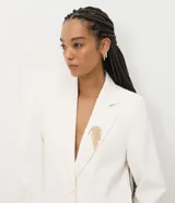 Blazer estructurado color blanco de la marca Cortelle, con solapa clásica, bolsillos frontales con solapa, mangas largas y cierre frontal de botones. Incluye un broche decorativo dorado con flecos de pedrería.