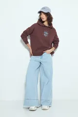 Canguro negro de calce relajado con capucha y cordones ajustables. Presenta un estampado gráfico frontal de un ojo con rayos de sol y la frase "STAY WEIRD".