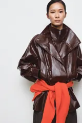 Chaqueta amplia de efecto piel en color marrón oscuro, con cuello alto, manga larga y bolsillos delanteros. Presenta cintura ajustable, forro interior y cierre frontal con cremallera y botones ocultos por solapa.