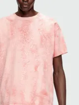 Remera de manga corta, cuello redondo y calce holgado, color rosa pálido con un efecto de teñido o estampado que simula grietas o craquelado en tonos más oscuros de rosa/naranja.