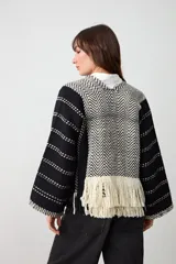 Chaqueta abierta de tejido de punto con diseño geométrico en blanco y negro. Presenta solapas con patrón de espiga y terminaciones con flecos largos en el frente. Posee mangas anchas con detalle de líneas horizontales punteadas.