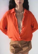 Camisa de corte oversize color naranja, con cuello camisero, cierre frontal de botones y mangas largas con puños abotonados. Presenta un acabado con efecto arrugado.