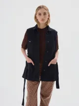 Chaleco de jean negro, con cuello camisero, cierre frontal de botones, dos bolsillos con solapa en el pecho y dos bolsillos de parche en la parte inferior. Incluye tiras ajustables en los laterales.