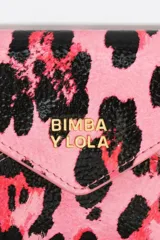 Monedero de gran tamaño con textura brillante y estructura rígida. Presenta un estampado animal print en tonos amarillo claro, negro y toques de azul. Cierre de botón a presión y logo metálico de Bimba y Lola en el frente.