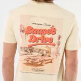 Remera de manga corta color beige, con cuello redondo y estampado frontal con la inscripción "Sunset Drive" y detalles tipográficos en el pecho.