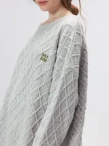 Sweater gris de punto con diseño de rombos, cuello redondo y mangas largas. Cuenta con puños largos acanalados con rayas horizontales en color verde militar y un pequeño logo bordado en el pecho.