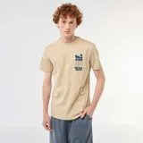 Camiseta de algodón color beige, de manga corta y cuello redondo, con diseño gráfico estampado en el pecho que incluye texto y olas estilizadas.