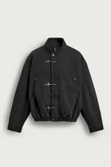 Cazadora de corte relaxed fit confeccionada en tejido de algodón de color negro. Cuenta con cuello subido, manga larga con puños abotonados, bolsillos de plastrón en la cadera y bajo elástico. Su diseño destaca por el cierre frontal de cremallera oculta con solapa decorada con alamares metálicos.
