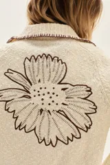Cardigan de punto color crudo con textura, cuello camisero con borde en contraste, mangas largas con puños acampanados y detalle de bordado floral en la espalda.